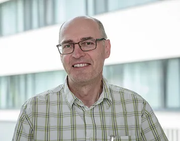 Ein lächelnder Mann mit Brille steht vor einem modernen Gebäude mit Glasfenstern. Er trägt ein kariertes Hemd und wirkt entspannt.