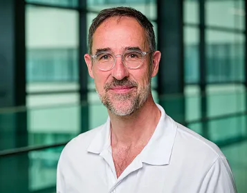 Porträt eines Mannes mit Brille und Bart in einem modernen Bürogebäude. Er trägt ein weißes, offenes Hemd und lächelt in die Kamera.