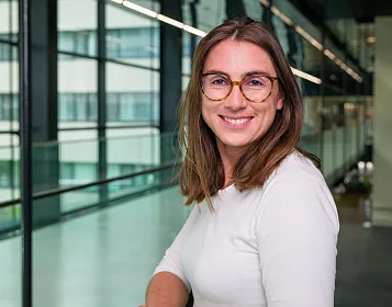Frau mit braunen Haaren und Brille lächelt in die Kamera. Sie trägt ein weißes Oberteil. Im Hintergrund moderne Bürogebäude mit Glaswänden.