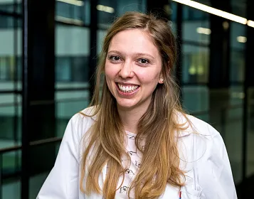 Junge Frau mit langen Haaren lächelt in die Kamera. Sie trägt einen weißen Kittel und steht in einem modernen Gebäude mit Glaswänden im Hintergrund.