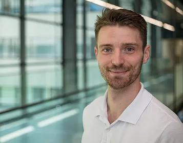 Mann mit kurzem braunem Haar und Bart, lächelt, trägt weißes Hemd. Im Hintergrund Glaswände mit Balkonblick. Helle und moderne Inneneinrichtung.