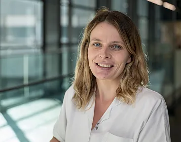 Eine lächelnde Person in einem weißen Kittel steht in einem modern gestalteten Innenraum mit viel Glas und Licht. Sie hat mittellanges, braunes Haar.