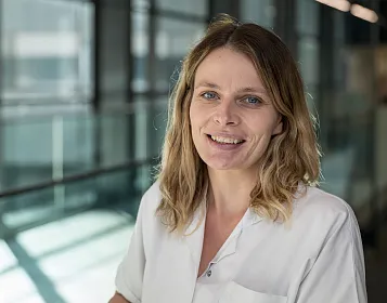Eine Frau mit blonden Haaren lächelt in die Kamera und trägt einen weißen Arztkittel. Sie steht vor einem Glasfenster, das den Hintergrund unscharf zeigt.