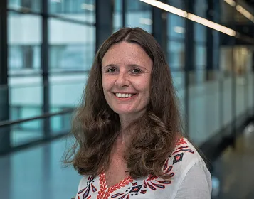 Eine Frau mit langen braunen Haaren trägt ein weißes Hemd mit rot-schwarzen Mustern. Sie lächelt und steht in einem modernen Innenraum mit großen Fenstern.
