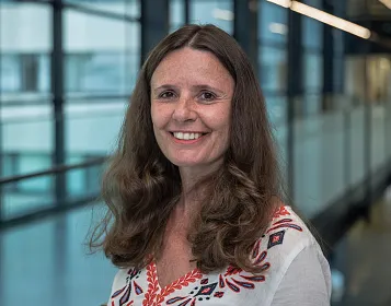 Eine Frau mit langen braunen Haaren lächelt in einem modernen Gebäude mit Glaswänden und einem beleuchteten Gang im Hintergrund.