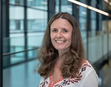 Frau mit langen braunen Haaren lächelt in einem modernen Bürogebäude mit großen Fenstern im Hintergrund. Sie trägt eine weiß-rote Bluse.