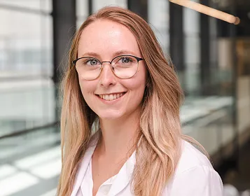 Eine lächelnde Person mit langen, blonden Haaren und Brille steht vor einem modernen Gebäude mit großen Fenstern. Sie trägt ein helles Oberteil.