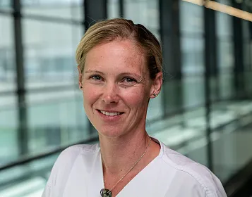 Porträt einer lächelnden Frau in weißem Oberteil mit hellem Haar. Sie steht vor einem Glasgebäude, das Licht reflektiert auf den Glaswänden.