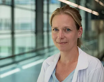 Eine Person in einem weißen Laborkittel steht in einem modernen Gebäude mit großen Glasfenstern. Sie hat blondes Haar und lächelt in die Kamera.