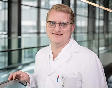 Ein Mann mit Brille und kurzen, blonden Haaren steht in einem modernen Gebäude. Er trägt einen weißen Kittel und lächelt in die Kamera.