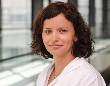 Eine Frau mit dunklem, welligem Haar steht in einem modernen Gebäude mit großen Fenstern. Sie trägt einen weißen Kittel und hat einen freundlichen Ausdruck.