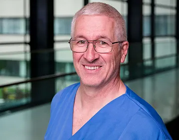 Ein lächelnder Mann mittleren Alters mit Brille trägt blaue medizinische Kleidung vor einem modernen, verglasten Gebäudekorridor.