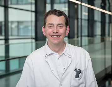 Ein lächelnder Mann in weißem Kittel steht in einem modernen Gebäude mit großen Fenstern im Hintergrund.