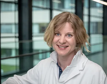 Eine Person mit kurzem blondem Haar trägt einen weißen Laborkittel und lächelt. Sie befindet sich in einem modernen Gebäude mit Glaswänden.