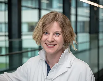 Eine Person mit schulterlangem, blonden Haar trägt einen weißen Laborkittel. Im Hintergrund sind moderne Bürofenster und ein glänzendes Geländer zu sehen.