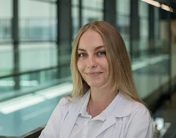 Eine Frau mit langen blonden Haaren und weißem Laborkittel lächelt in die Kamera. Sie steht in einem modernen, lichtdurchfluteten Gebäude.