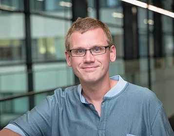 Mann mit Brille und kurzem Haar, trägt ein blaues Hemd. Er lächelt leicht und steht in einem modernen Gebäude mit Glasfronten und Bürobeleuchtung.