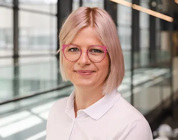 Frau mit blonden Haaren und pinken Brillen in weißer Bluse lächelt vor verglaster Innenarchitektur. Die Umgebung wirkt modern und hell ausgeleuchtet.