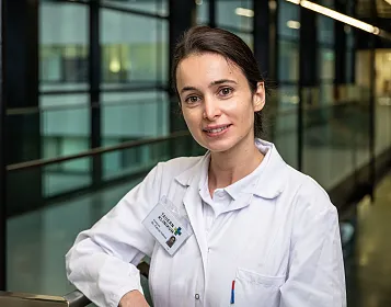 Ärztin in weißem Kittel lächelt in modernen Klinikfluren. Sie steht leicht gelehnt, trägt ein Namensschild und hat braune Haare, die zusammengebunden sind.