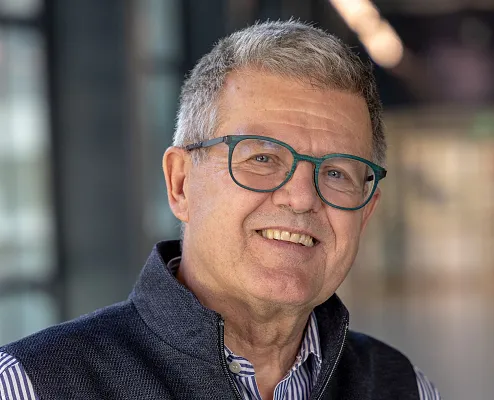 Ein lächelnder älterer Mann mit Brille trägt ein gestreiftes Hemd und eine graue Weste. Der Hintergrund ist verschwommen und vermittelt eine klassische Innenraumszene.