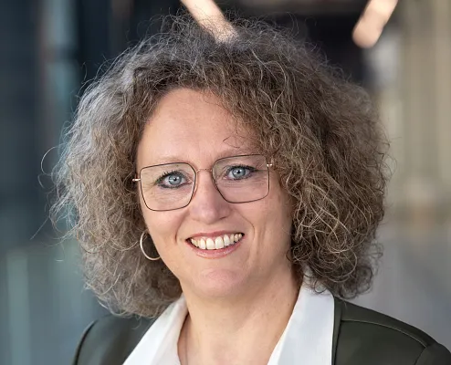 Porträt einer lächelnden Person mit lockigem Haar, Brille und einem dunklen Blazer über einer weißen Bluse. Der Hintergrund ist unscharf.