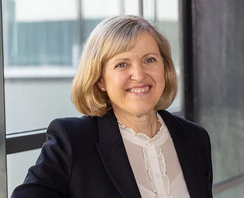 Eine lächelnde Person in einem schwarzen Blazer und weißer Bluse steht vor einem Fenster. Der Hintergrund ist unscharf und neutral, die Stimmung professionell.