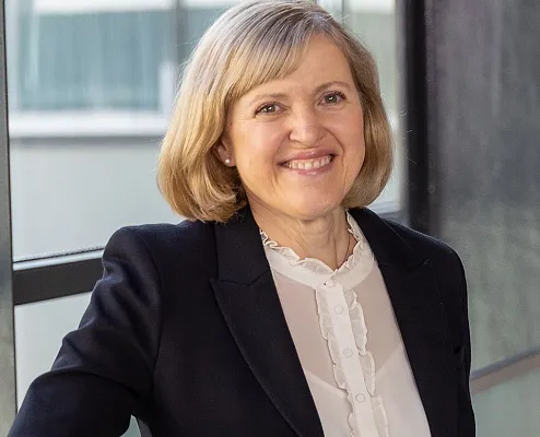 Eine lächelnde Person in einem schwarzen Blazer und weißer Bluse steht vor einem Fenster. Der Hintergrund ist unscharf und neutral, die Stimmung professionell.