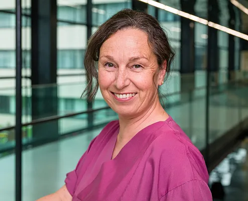 Eine Frau in pinker Arbeitskleidung lächelt in die Kamera. Sie steht in einem modernen, verglasten Gebäude mit Fensterfront im Hintergrund.