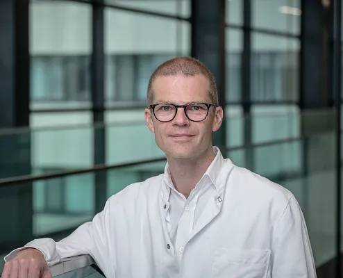 Ein Mann mit Brille trägt einen weißen Laborkittel und lächelt leicht. Er steht in einem modernen Gebäude mit großen Fenstern und einer Glasbalustrade.