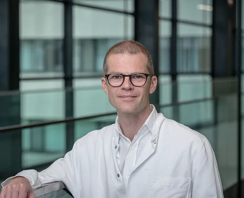 A person in a white lab coat with glasses, standing in a modern glass building, suggesting a scientific environment.
