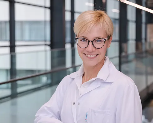 Eine lächelnde Person in einem weißen Laborkittel steht in einem modernen Gebäude mit Glaswänden. Sie trägt eine Brille und hat kurzes blondes Haar.