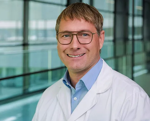 Porträt eines lächelnden Mannes in einem weißen Kittel und blauer Hemd, mit Brille, in einem modernen, hellen Gebäude mit Glaswänden im Hintergrund.