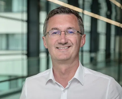 Ein Mann mit Brille und kurzem Haar lächelt freundlich in die Kamera. Er trägt ein weißes Hemd. Der Hintergrund zeigt ein modernes Bürogebäude.