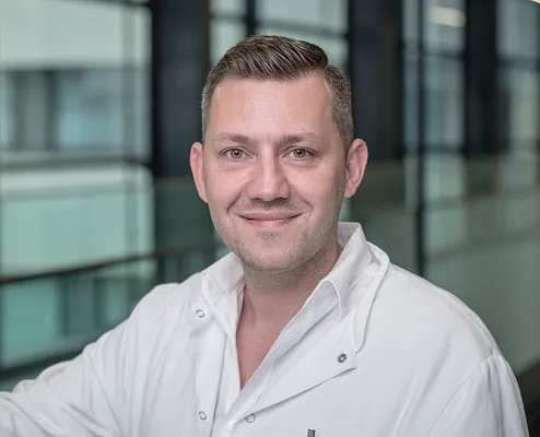 Ein Mann in einem weißen Laborkittel lächelt in die Kamera. Der Hintergrund zeigt ein modernes Gebäude mit großen Glasfenstern, die viel Licht zulassen.