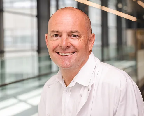 Ein lächelnder Mann in einem weißen Kittel steht in einem modernen, lichtdurchfluteten Gebäude mit großen Fenstern im Hintergrund.