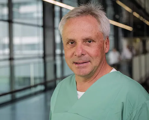 Ein Mann in grüner medizinischer Kleidung steht in einem modernen Gebäude mit Glaswänden. Er hat graues Haar und schaut direkt in die Kamera.