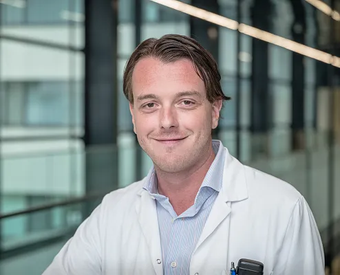 Ein Mann in einem weißen Kittel steht lächelnd in einem modernen Gebäude mit Glasfronten. Er trägt ein helles Hemd und hat kurze, dunkelblonde Haare.