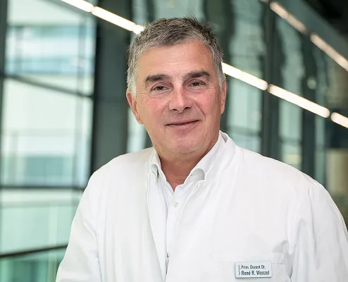 Ein lächelnder Mann mittleren Alters in einem weißen Arztkittel steht in einem modernen Gebäude mit großen Fenstern.