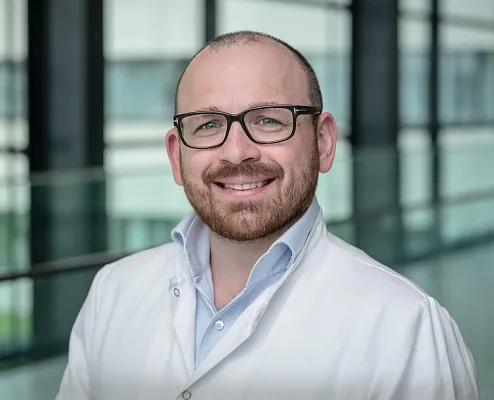 Ein lächelnder Arzt mit Brille und Bart steht in einem modernen, lichtdurchfluteten Gebäude. Er trägt einen weißen Kittel vor Glasfassaden.