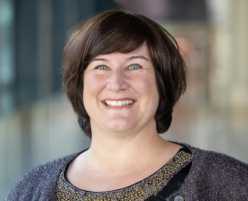 Eine lächelnde Person trägt einen glitzernden Schal und ein Oberteil mit dekorativem Kragen. Der Hintergrund ist verschwommen und zeigt eine neutrale Umgebung.