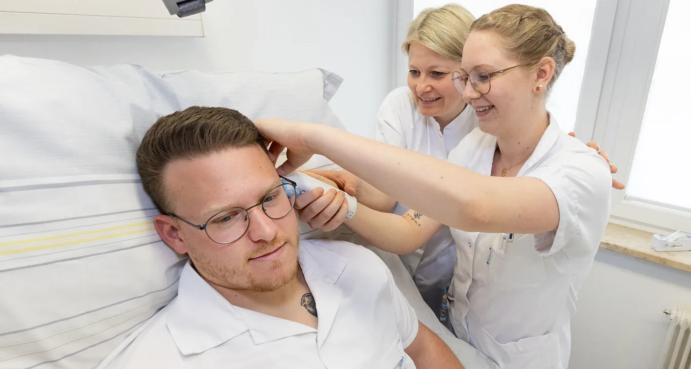Zwei Pflegerinnen in weißer Dienstkleidung versorgen einen männlichen Patienten im Krankenbett, indem sie seine Haare schneiden und pflegen.