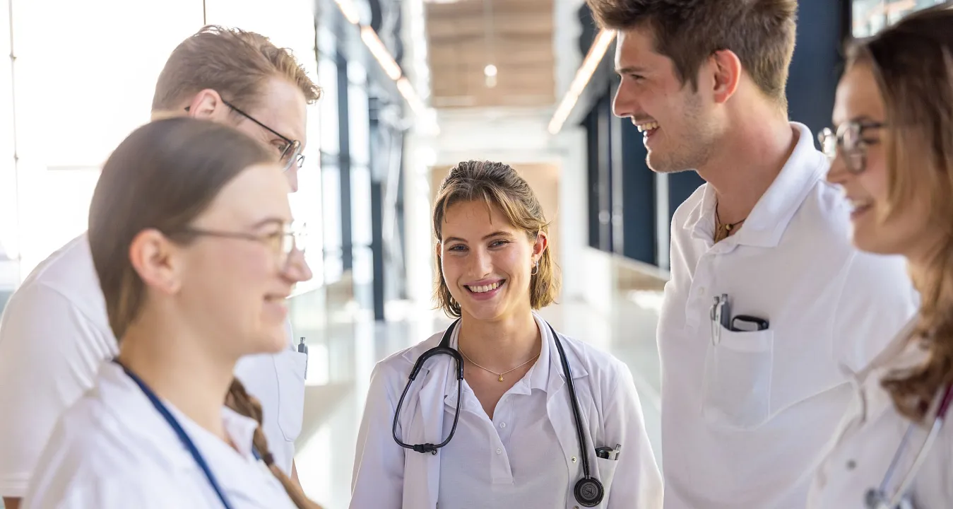 Eine Gruppe von fünf medizinischen Fachkräften in weißen Kitteln unterhält sich freundlich in einem modernen Klinikflur. Sie tragen Stethoskope.