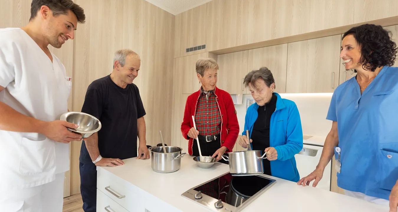 Gruppe von älteren Menschen und Betreuer:Innen bereiten in moderner Küche gemeinsam Speisen zu. Alle wirken engagiert und haben Kochutensilien in den Händen.