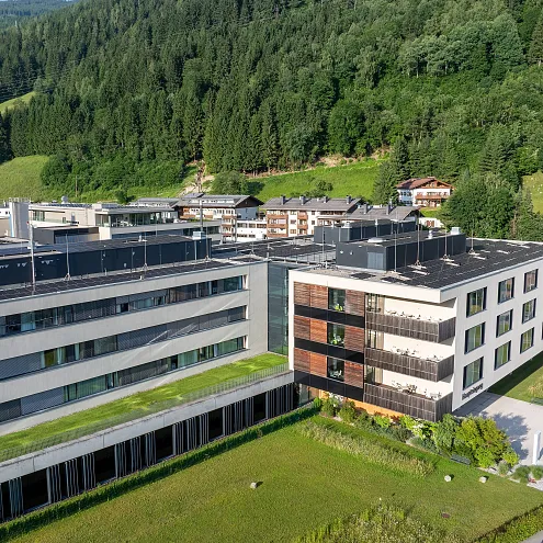 Modern building in a mountain landscape, featuring flat roofs, large windows, and greenery. Surrounded by hills and trees under a clear sky.