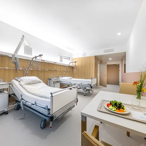 Ein modernes Krankenhauszimmer mit zwei leeren Krankenbetten, gut beleuchtet. Ein Tisch mit einer Servierplatte und einem Teller Salat steht im Vordergrund.