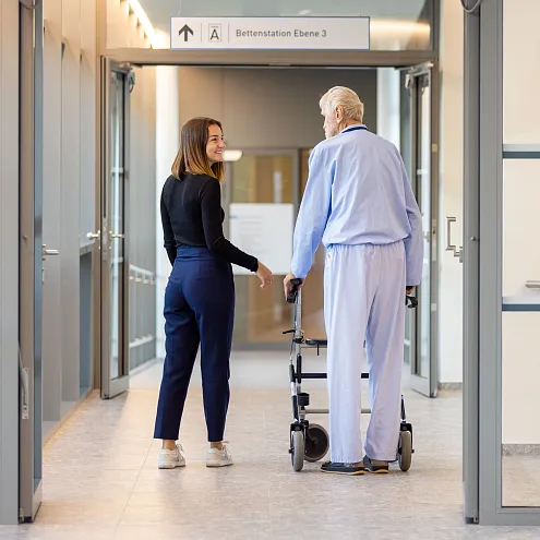 Eine Frau begleitet einen älteren Mann im Krankenhausflur. Der Mann benutzt einen Rollator, und beide lächeln. Gemeinschaft und Unterstützung sind erkennbar.