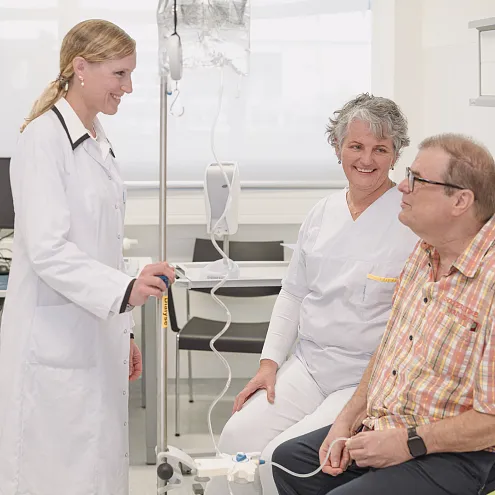 Eine Ärzt:in spricht mit einem sitzenden Patient:innen, während eine Pfleger:in daneben lächelt. Im Hintergrund sind medizinische Geräte und Bürostühle zu sehen.