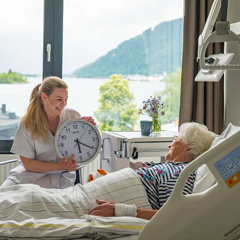 Eine Pflegekraft zeigt einer älteren Frau, die im Krankenhausbett liegt, eine Wanduhr. Im Hintergrund ist ein Fenster mit Seeblick zu sehen.