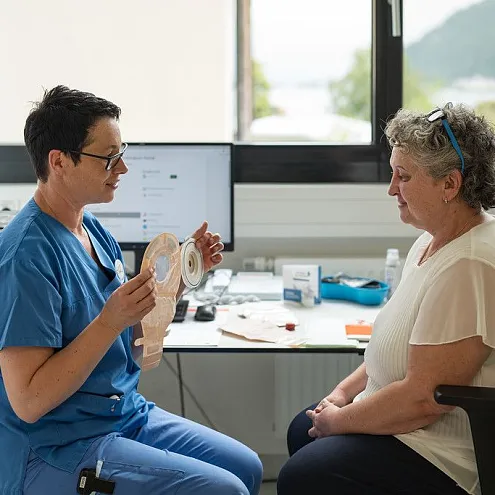 Gesundheitsfachkraft in blauem Kittel zeigt einer älteren Frau medizinisches Hilfsmittel. Beide sitzen in einem hellen Raum mit einem Fenster im Hintergrund.
