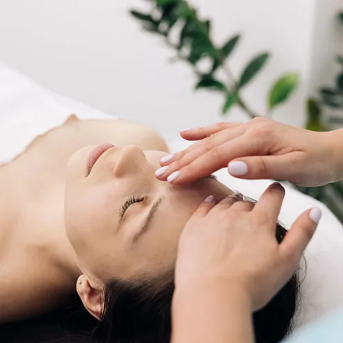 Eine Person liegt entspannt auf einer Massageliege, während eine andere Person sanft die Gesichtsmassage durchführt. Pflanzen sind im Hintergrund sichtbar.